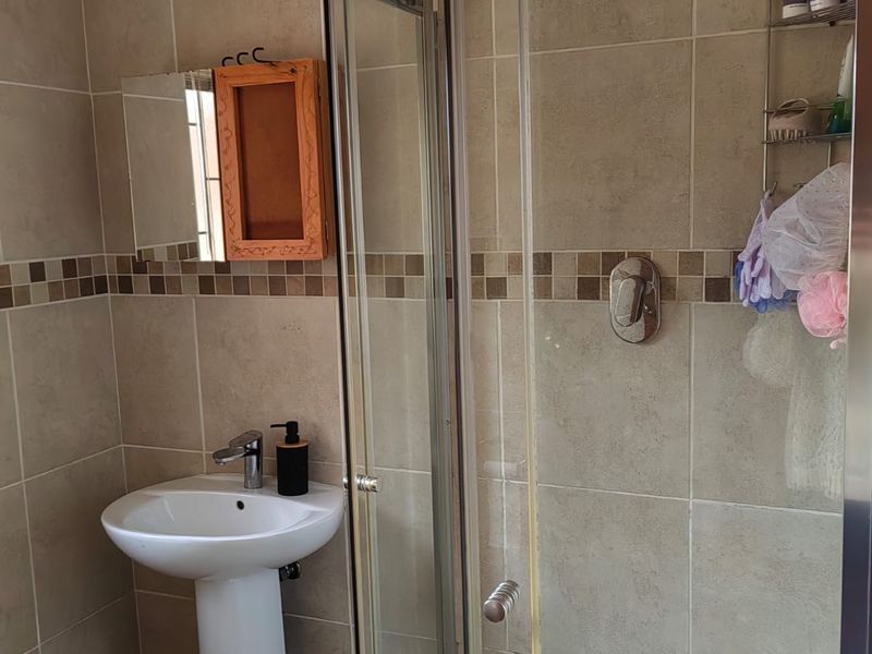 A functional bathroom featuring a pedestal basin, a glass-enclosed shower with a rain shower head, and neutral ceramic wall tiling with a mosaic accent.