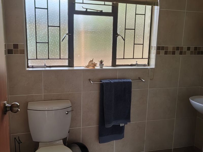 A functional bathroom featuring neutral ceramic wall and floor tiles, a standard toilet, pedestal basin, and a window fitted with security bars.