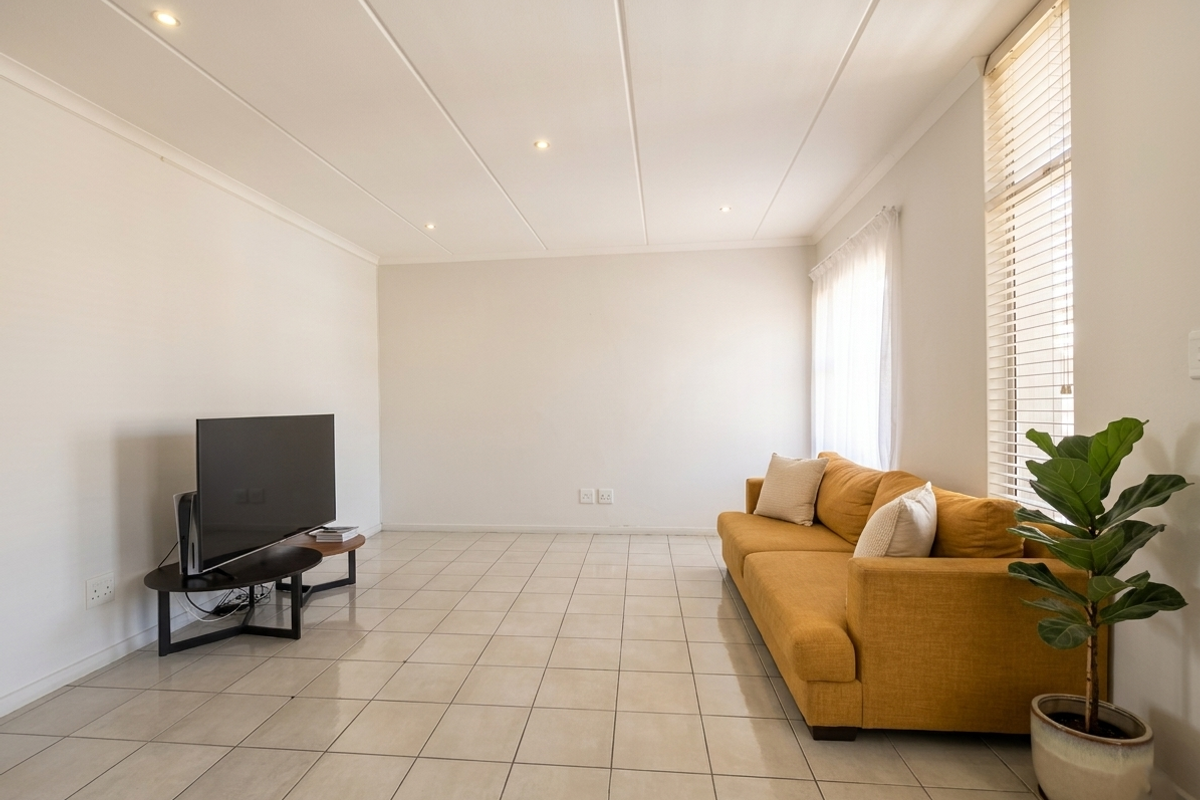 A bright and spacious living room featuring neutral tiled flooring, recessed ceiling lighting, and large windows fitted with venetian blinds.