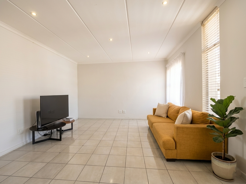 A bright and spacious living room featuring neutral tiled flooring, recessed ceiling lighting, and large windows fitted with venetian blinds.