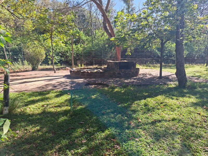 A spacious garden area featuring a stone-built braai facility, mature trees, and perimeter wire fencing on a small holding property.