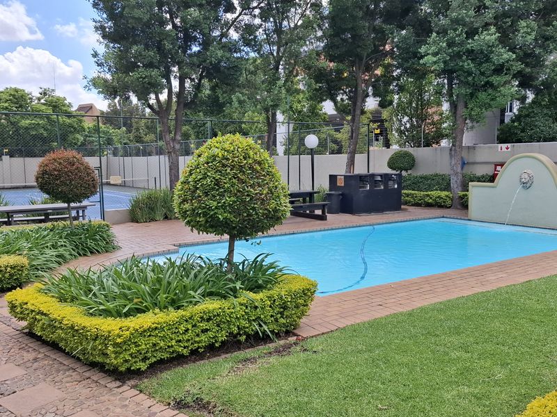 Well-maintained communal pool area featuring a water fountain, paved surround, landscaped gardens, and an adjacent tennis court within the complex.
