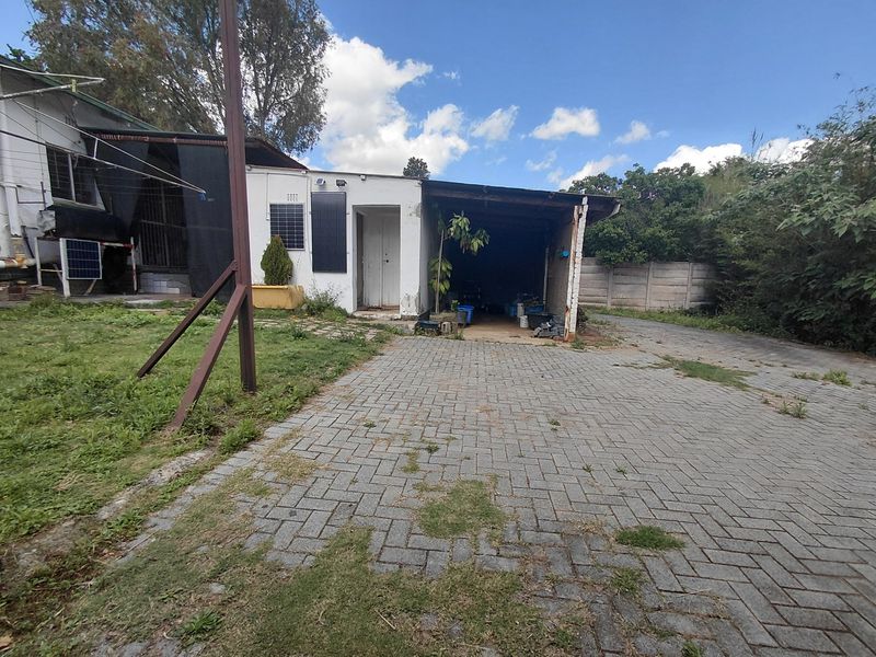 Rear exterior view showing a paved driveway, open carport structure, and boundary wall with signs of wear and maintenance requirements.