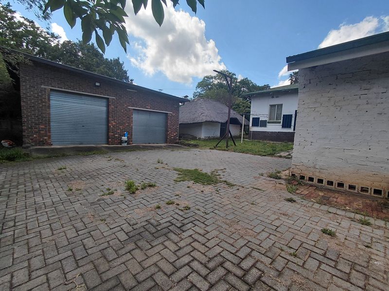 Rear courtyard area featuring a double garage with roller doors, paved driveway, and a secondary thatched structure in a spacious outdoor setting.