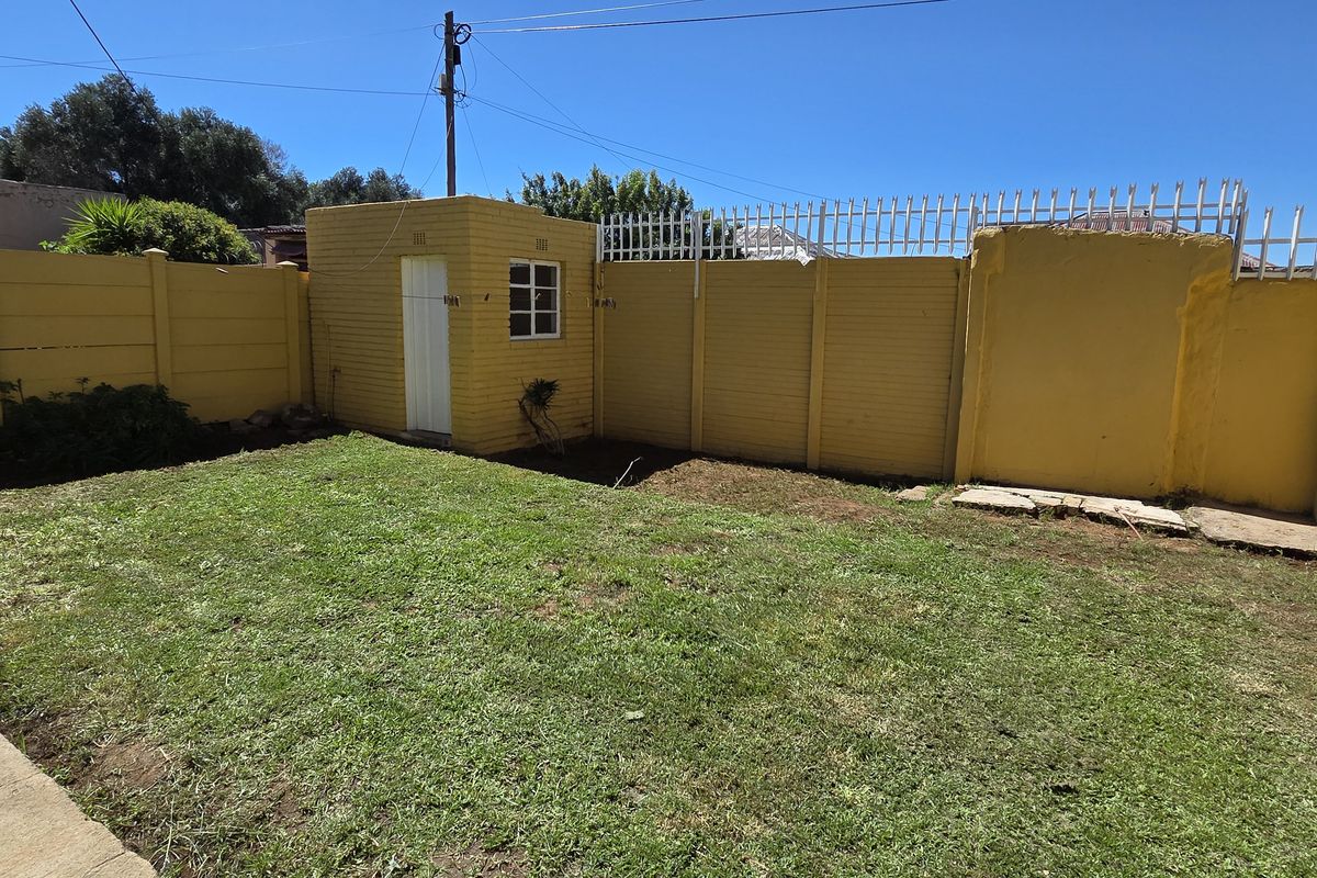A rear garden area featuring a lawn, concrete paving, and a small outbuilding enclosed by a yellow precast concrete boundary wall with security spikes.