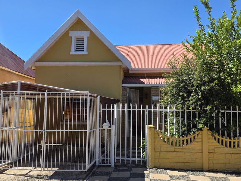 Front elevation of a house featuring a gabled corrugated metal roof, secure perimeter fencing, and a covered carport area.