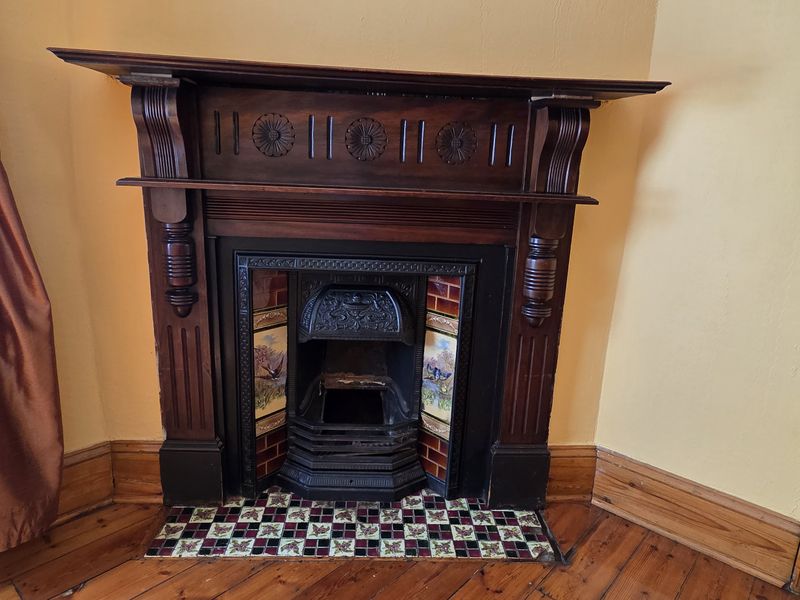 A character-filled living room featuring an ornate timber fireplace mantel with decorative cast iron insert and original tiled hearth, set against timber flooring.