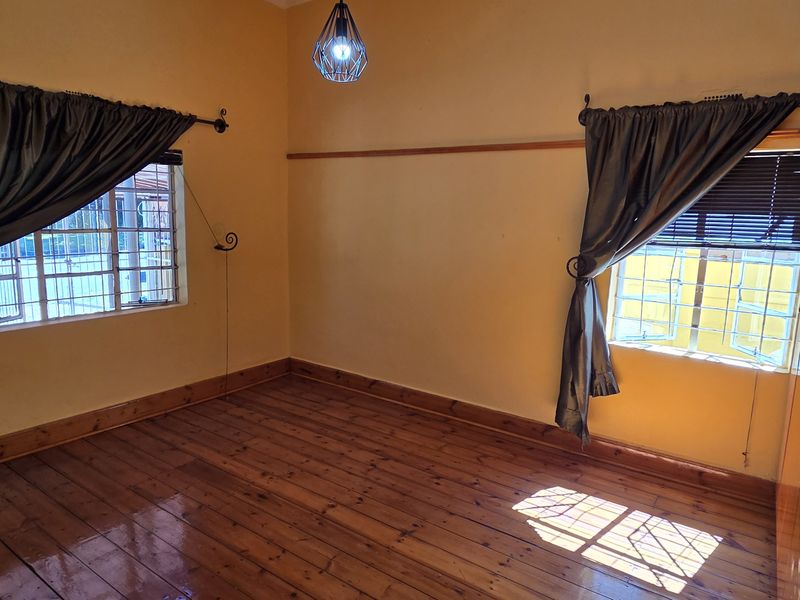 A bright, empty bedroom featuring polished hardwood flooring, built-in wooden cupboards, and security bars on the windows.