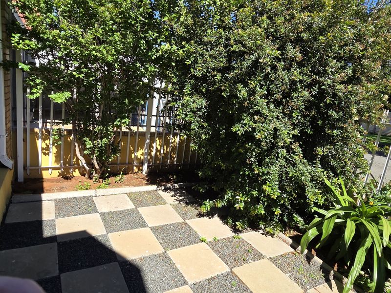 A small outdoor patio area featuring checkerboard paving, enclosed by metal security fencing and established garden greenery.