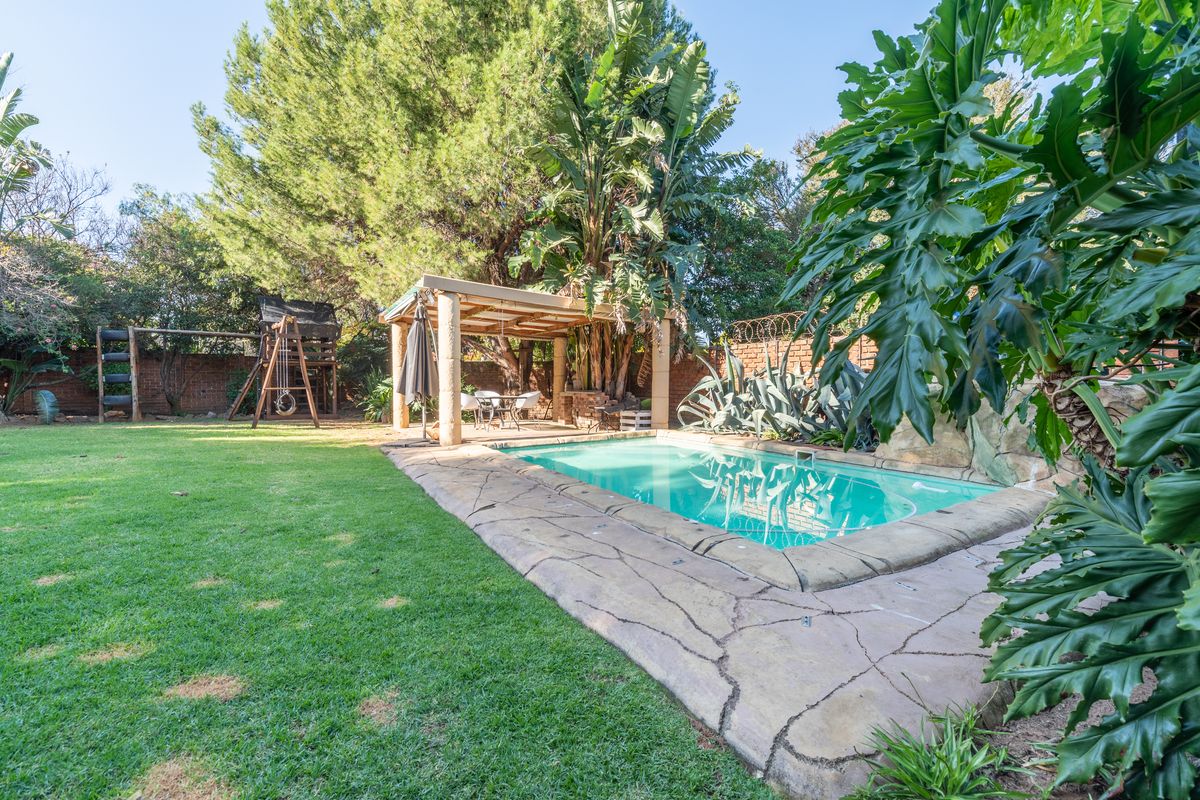 A private backyard featuring a swimming pool with stone coping, a covered patio area, and a well-maintained lawn surrounded by mature trees and shrubs.