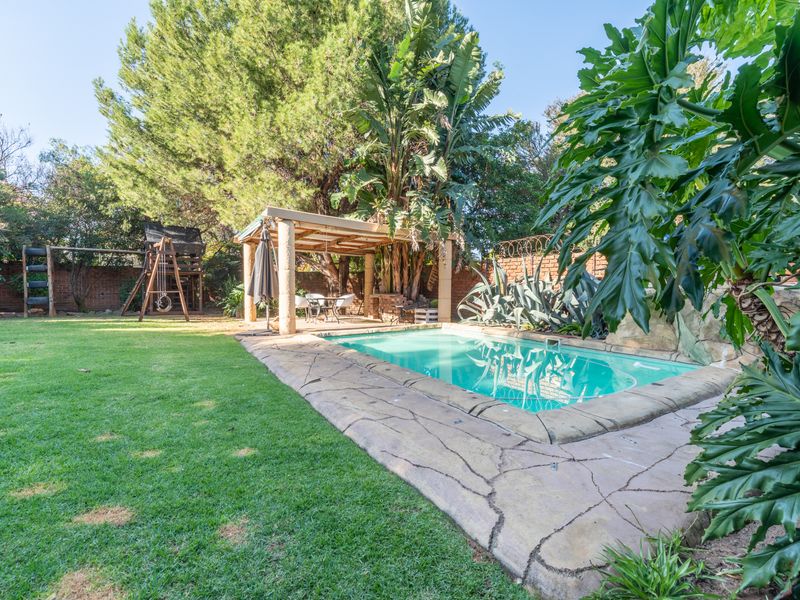 A private backyard featuring a swimming pool with stone coping, a covered patio area, and a well-maintained lawn surrounded by mature trees and shrubs.
