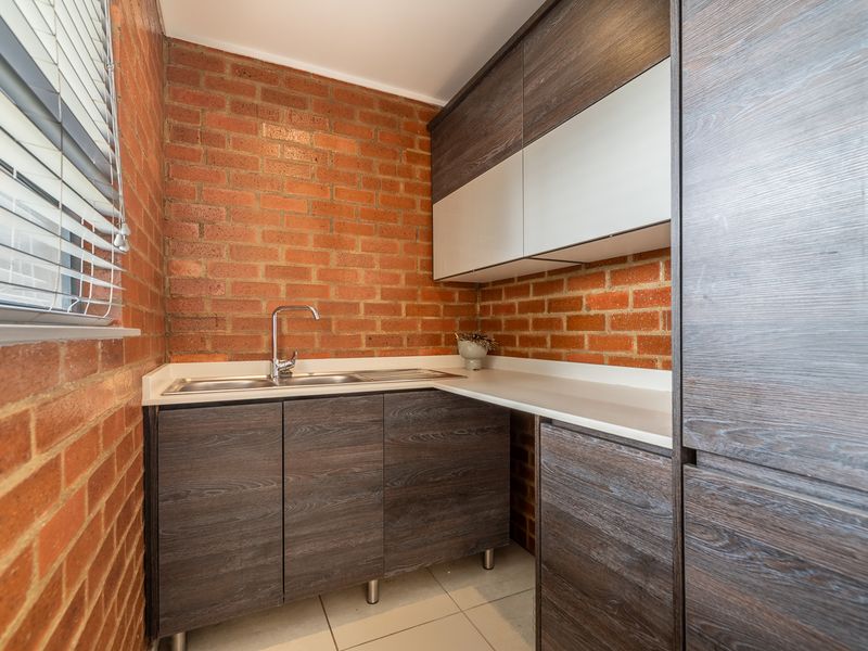 A compact, modern scullery featuring exposed brick walls, dark wood-grain cabinetry, stone-look countertops, and a stainless steel sink.