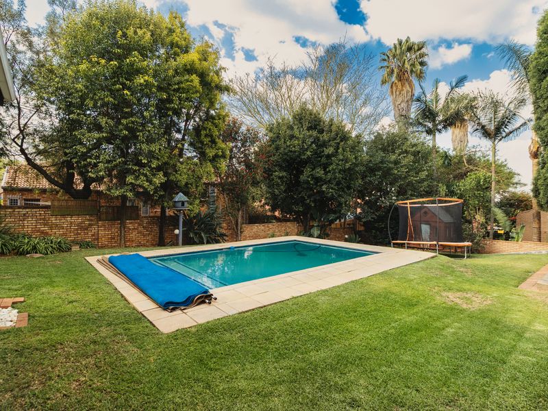 A well-maintained outdoor pool area featuring a clear swimming pool with surrounding paved coping and a lush, established garden with brick boundary walls.