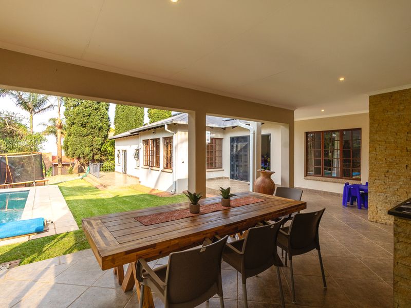A spacious covered patio featuring tiled flooring, recessed lighting, and a stone-clad feature wall, overlooking the garden and swimming pool area.