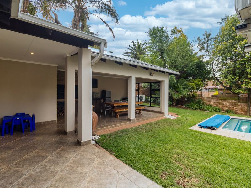 Spacious covered patio area featuring tiled flooring and a built-in braai, overlooking a private garden and swimming pool.