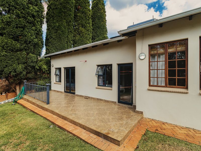 A well-maintained rear exterior view featuring a tiled patio, metal balustrade, brick paving, and timber-framed windows set against a landscaped garden.