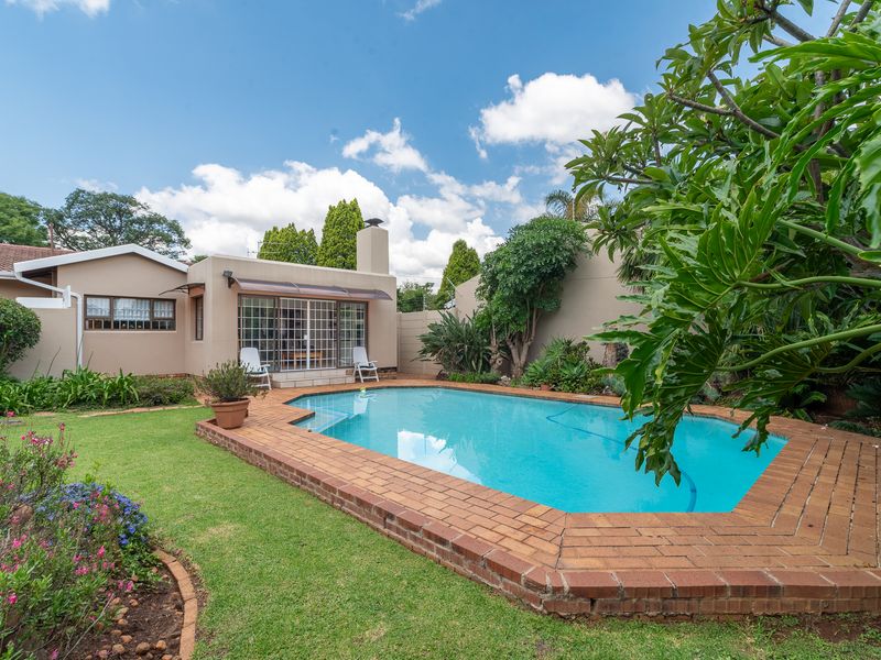 A well-maintained backyard featuring a swimming pool with brick coping, an outdoor fireplace, and a landscaped garden area.