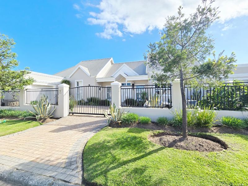 A well-maintained front facade featuring a paved driveway, secure wrought iron gate, perimeter wall, and neat landscaping under a metal roof.