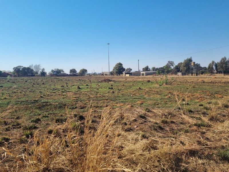 A wide-angle view of a vacant residential plot featuring dry grass, a water tank, and utility infrastructure in the distance.