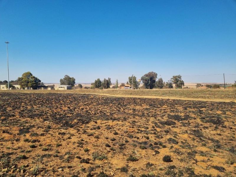 A wide-angle view of a vacant residential plot featuring scorched earth, distant trees, and visible utility infrastructure under a clear blue sky.