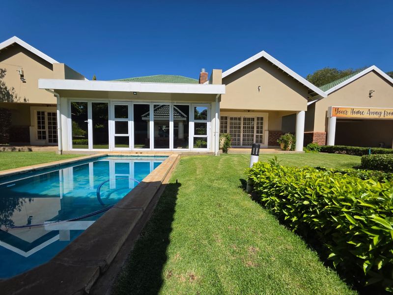 A well-maintained guest house exterior featuring a swimming pool, manicured lawn, and a covered patio area with large sliding glass doors.