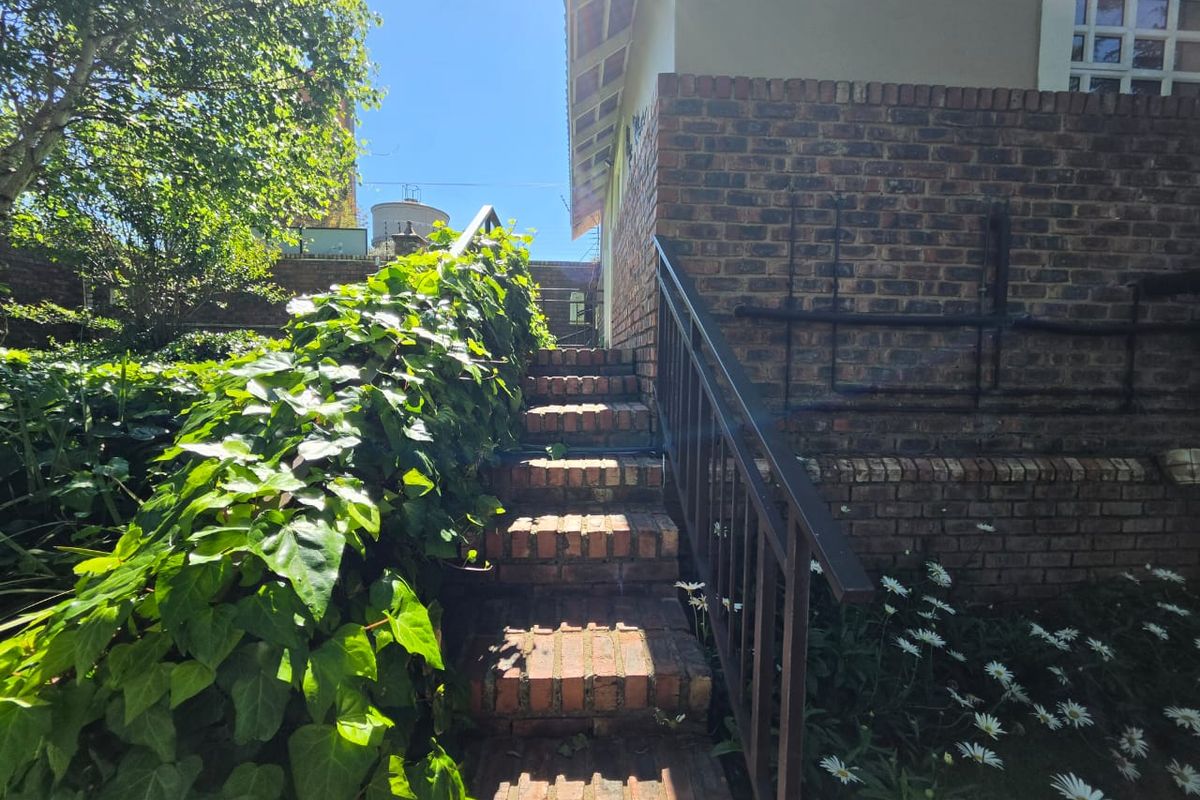 An outdoor brick staircase with a metal handrail leading up along the side of a brick building in a garden setting.