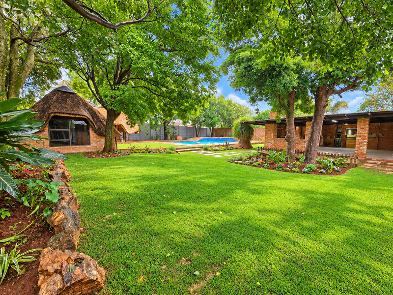 A beautifully landscaped garden featuring a lush lawn, mature trees, a swimming pool, and a charming thatched-roof structure with a brick patio area.
