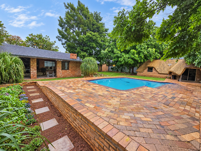 Spacious outdoor entertainment area featuring a swimming pool, extensive brick paving, and a charming thatched-roof outbuilding set within a mature garden.