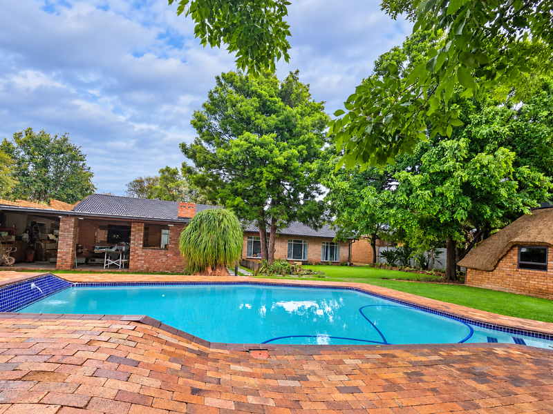 A well-maintained outdoor pool area featuring brick paving, a covered patio, and a charming thatched-roof outbuilding set within a lush garden.