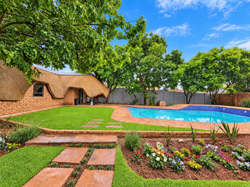 A well-maintained outdoor pool area featuring a swimming pool, paved surround, landscaped garden, and a distinctive thatched-roof brick structure.