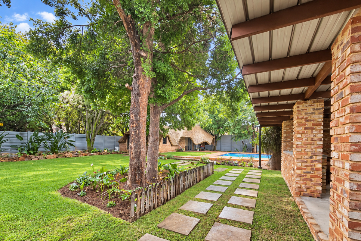 A spacious rear garden featuring a covered brick patio, a well-maintained lawn, mature trees, a paved walkway, and a swimming pool in the background.