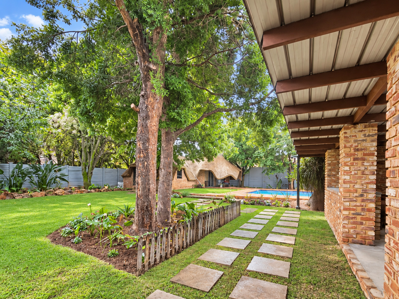 A spacious rear garden featuring a covered brick patio, a well-maintained lawn, mature trees, a paved walkway, and a swimming pool in the background.