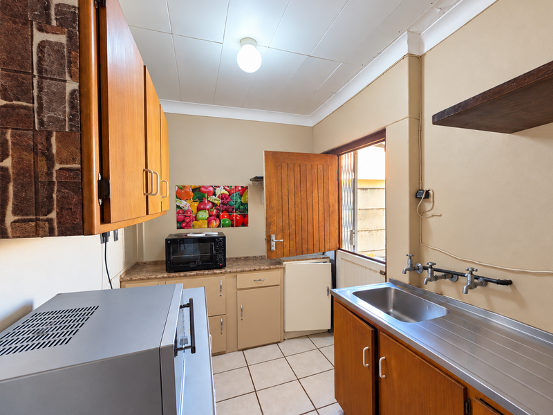 A functional scullery area featuring built-in wooden cabinetry, a stainless steel sink, tiled flooring, and direct access to the exterior.
