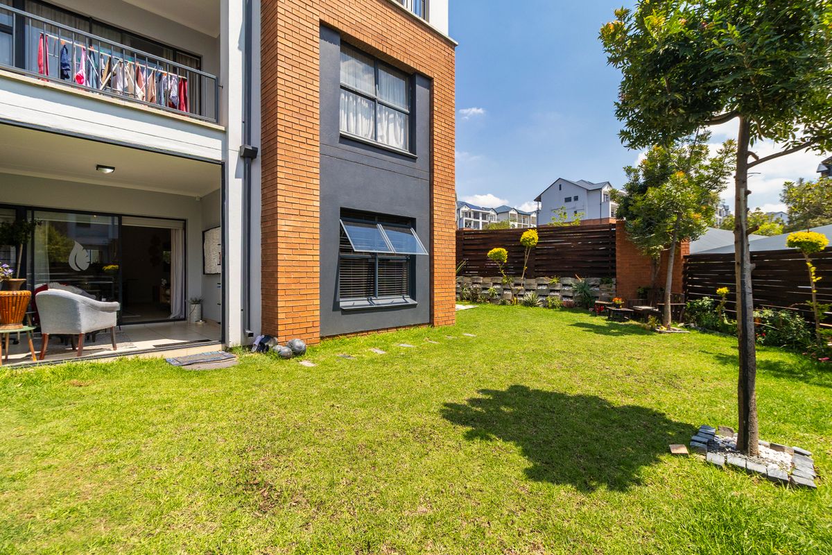 A well-maintained rear exterior view of an apartment building featuring a private garden, brick facade, and a ground-floor patio with sliding door access.