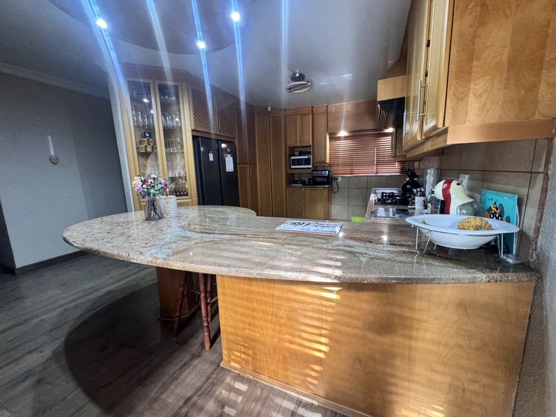 A spacious kitchen featuring extensive wooden cabinetry, a large granite breakfast bar, built-in cooking appliances, and modern recessed lighting.