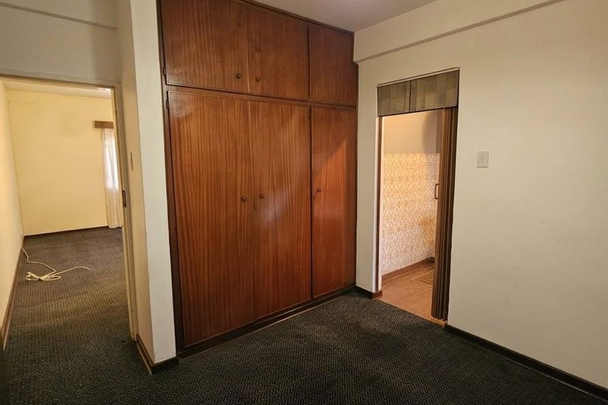An empty bedroom featuring built-in wooden wardrobes, carpet flooring, and a pendant light fixture, with an adjacent doorway leading to another room.