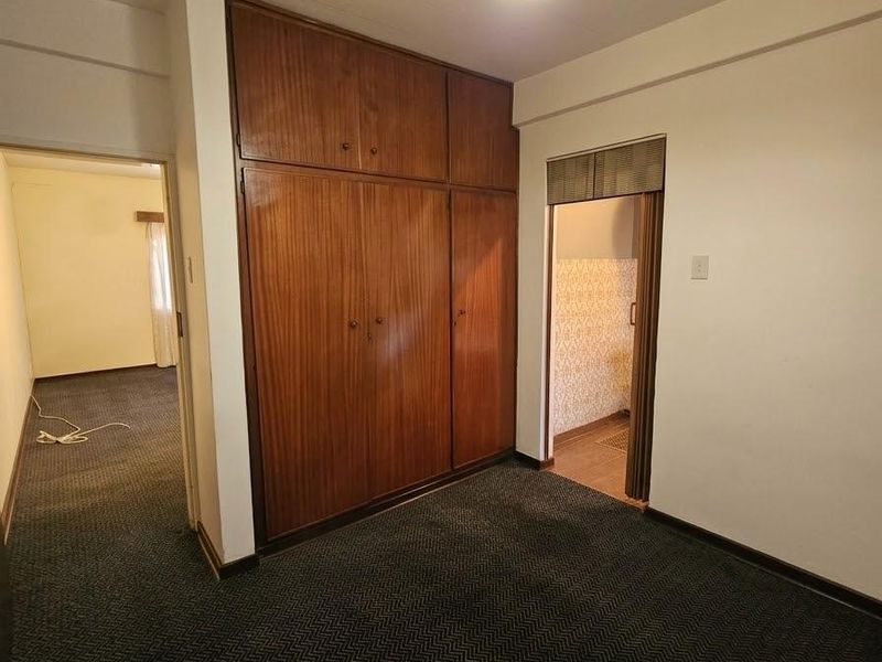 An empty bedroom featuring built-in wooden wardrobes, carpet flooring, and a pendant light fixture, with an adjacent doorway leading to another room.