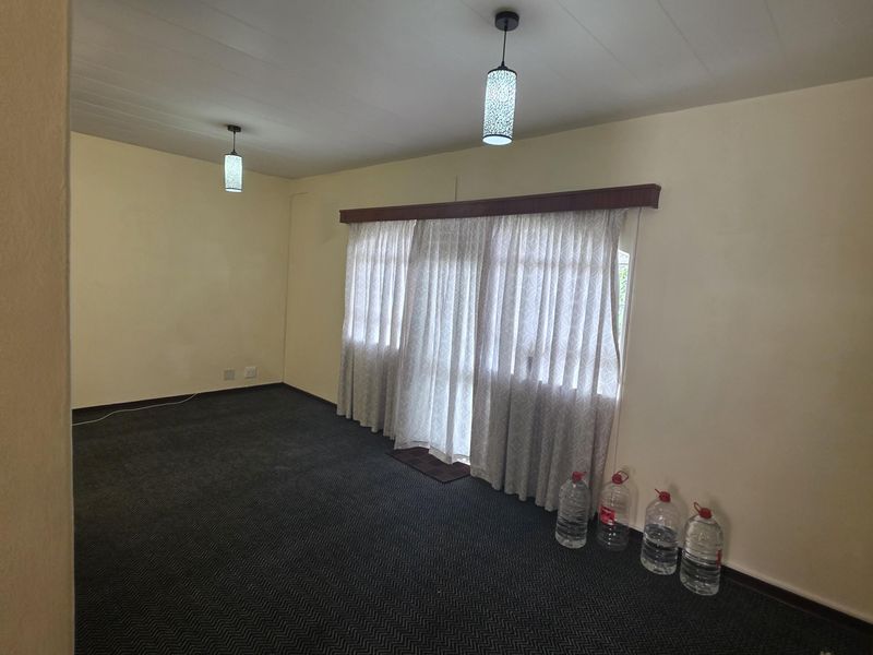 A spacious, empty living room featuring neutral walls, dark patterned carpeting, and dual pendant light fixtures with a large window covered by curtains.