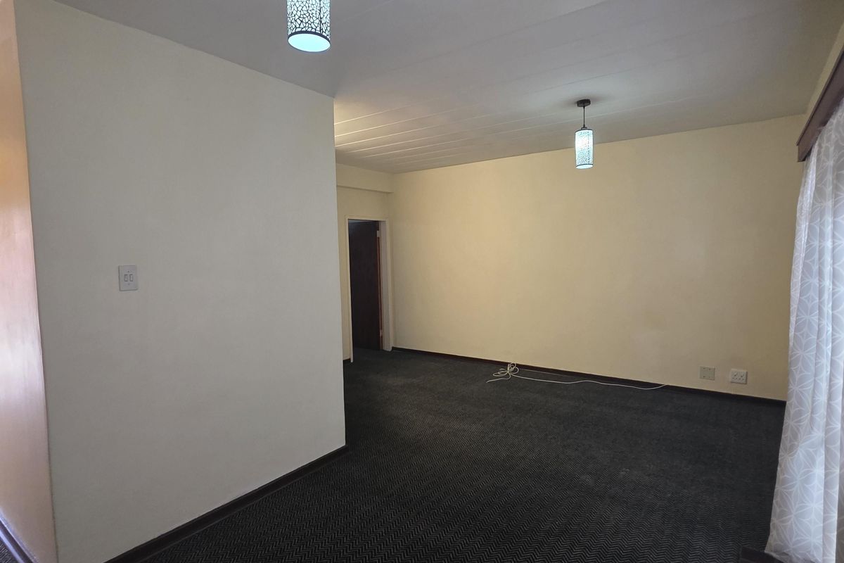 A spacious, empty living area featuring neutral walls, dark patterned carpeting, and modern pendant lighting fixtures.