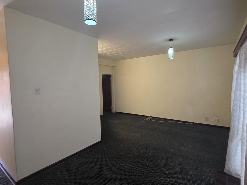 A spacious, empty living area featuring neutral walls, dark patterned carpeting, and modern pendant lighting fixtures.