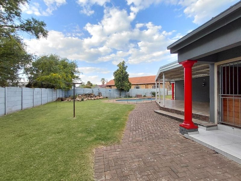 Spacious rear garden featuring a paved patio area, covered veranda with decorative columns, and a swimming pool enclosed by a safety fence.