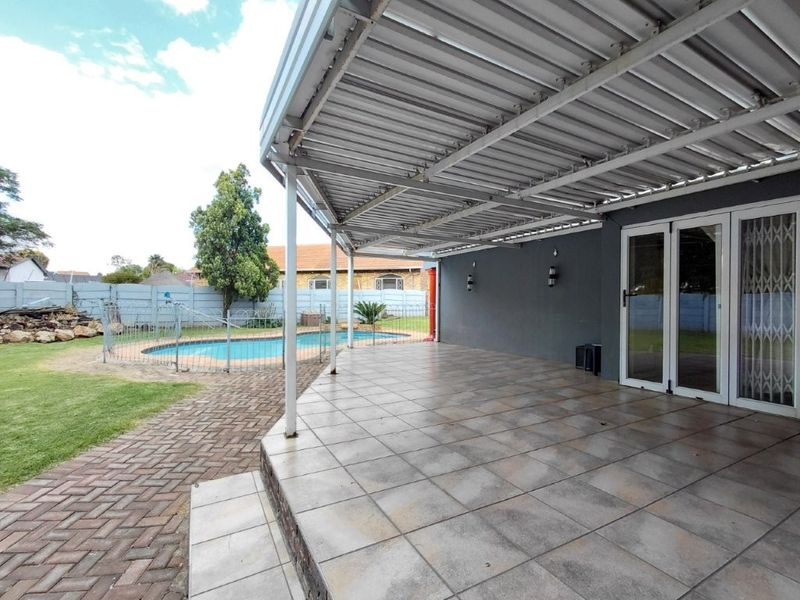 A spacious covered patio with tiled flooring, overlooking a private swimming pool and garden area, featuring direct access to the home via sliding doors.