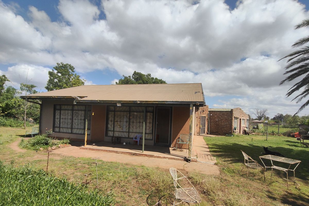 Rear view of a single-story brick house featuring a covered patio area, corrugated metal roofing, and security bars on windows, set within a large garden.