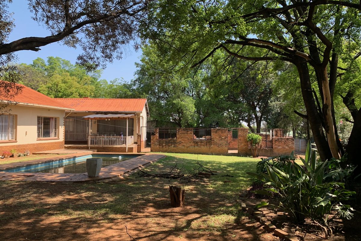 Rear garden view featuring a swimming pool, paved surround, covered patio, and established trees, set against a brick boundary wall.