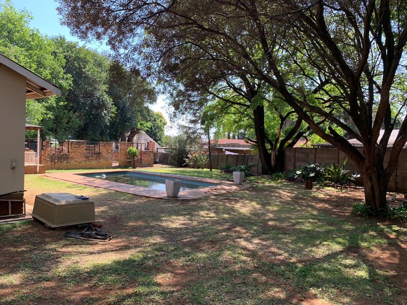 A spacious backyard featuring a swimming pool with brick coping, surrounded by mature trees and a mix of brick and concrete boundary walls.