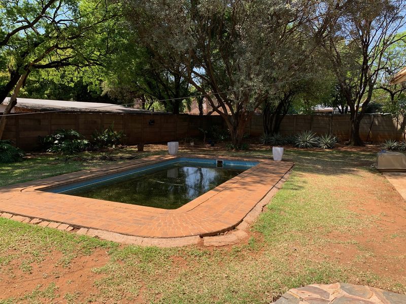 Outdoor pool area featuring a rectangular swimming pool with brick coping, surrounded by a lawn and mature trees, enclosed by a precast boundary wall.