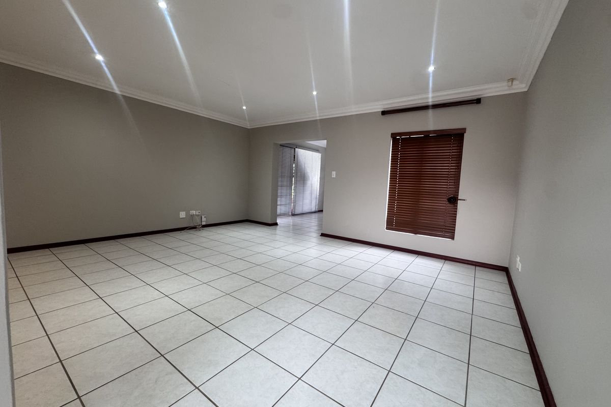 A spacious, empty living room featuring neutral tiled flooring, recessed downlighting, and venetian blinds, offering a clean and versatile blank canvas.