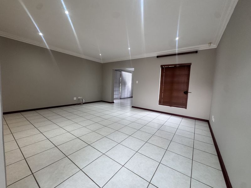 A spacious, empty living room featuring neutral tiled flooring, recessed downlighting, and venetian blinds, offering a clean and versatile blank canvas.