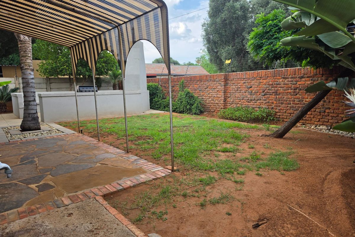Rear garden area featuring a paved patio, built-in braai, and a shade structure, enclosed by a brick boundary wall.