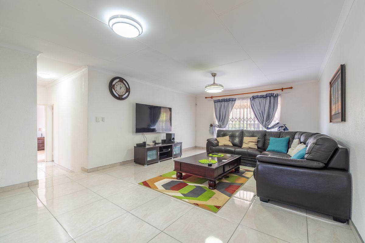 A spacious and bright living room featuring neutral tiled flooring, clean white walls, and a large window fitted with a security gate.